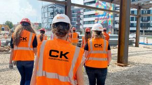 CK Construction crew members wearing branded orange high-visibility vests and white safety helmets walk the grounds of a new modular office construction site in Columbus, Ohio, with steel structural framing and a modern mixed-use building visible in the background.